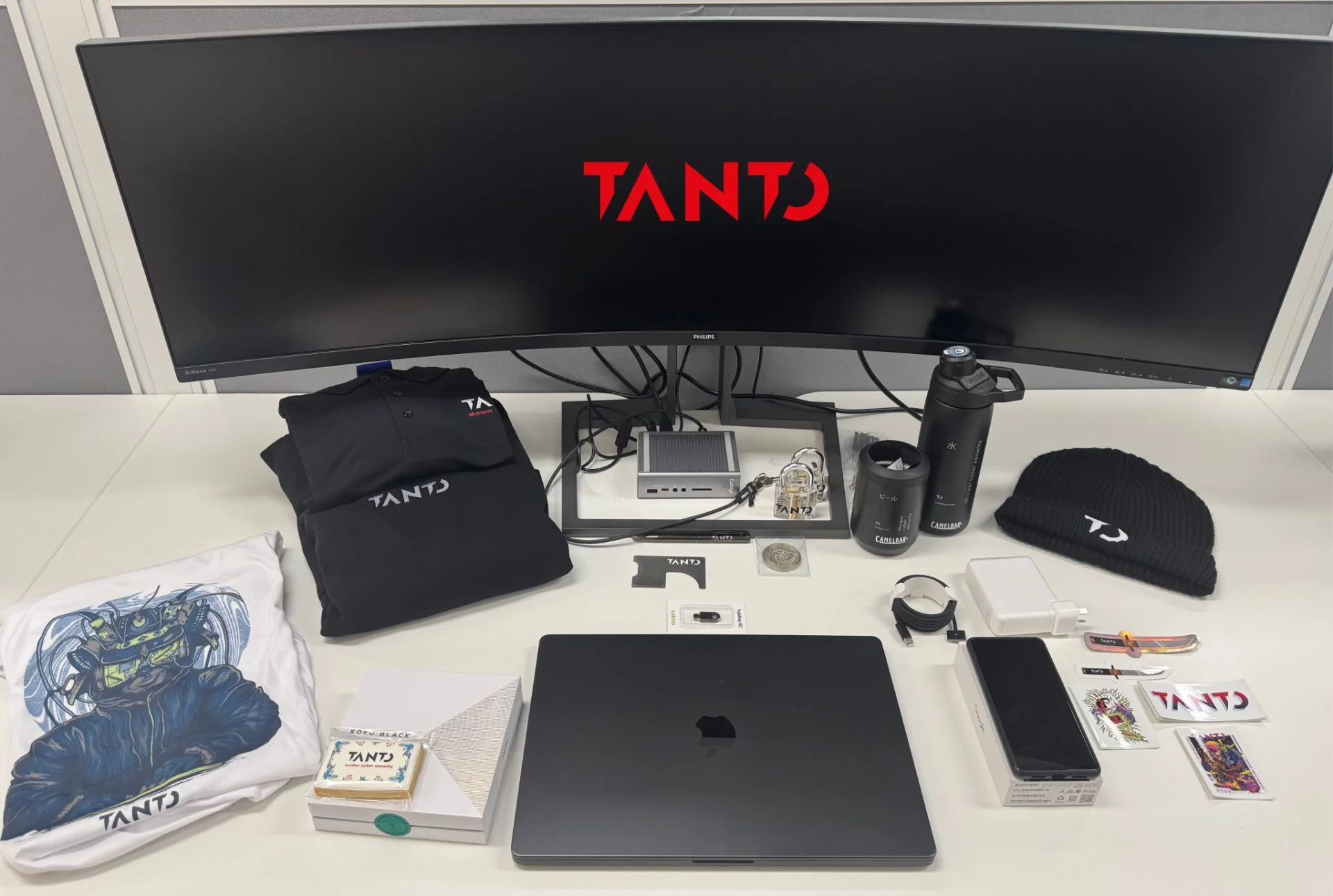 Items laid out on a desk including a MacBook, iPhone, t-shirt, polo shirt, YubiKey, challenge coin, water bottle, beanie and stickers